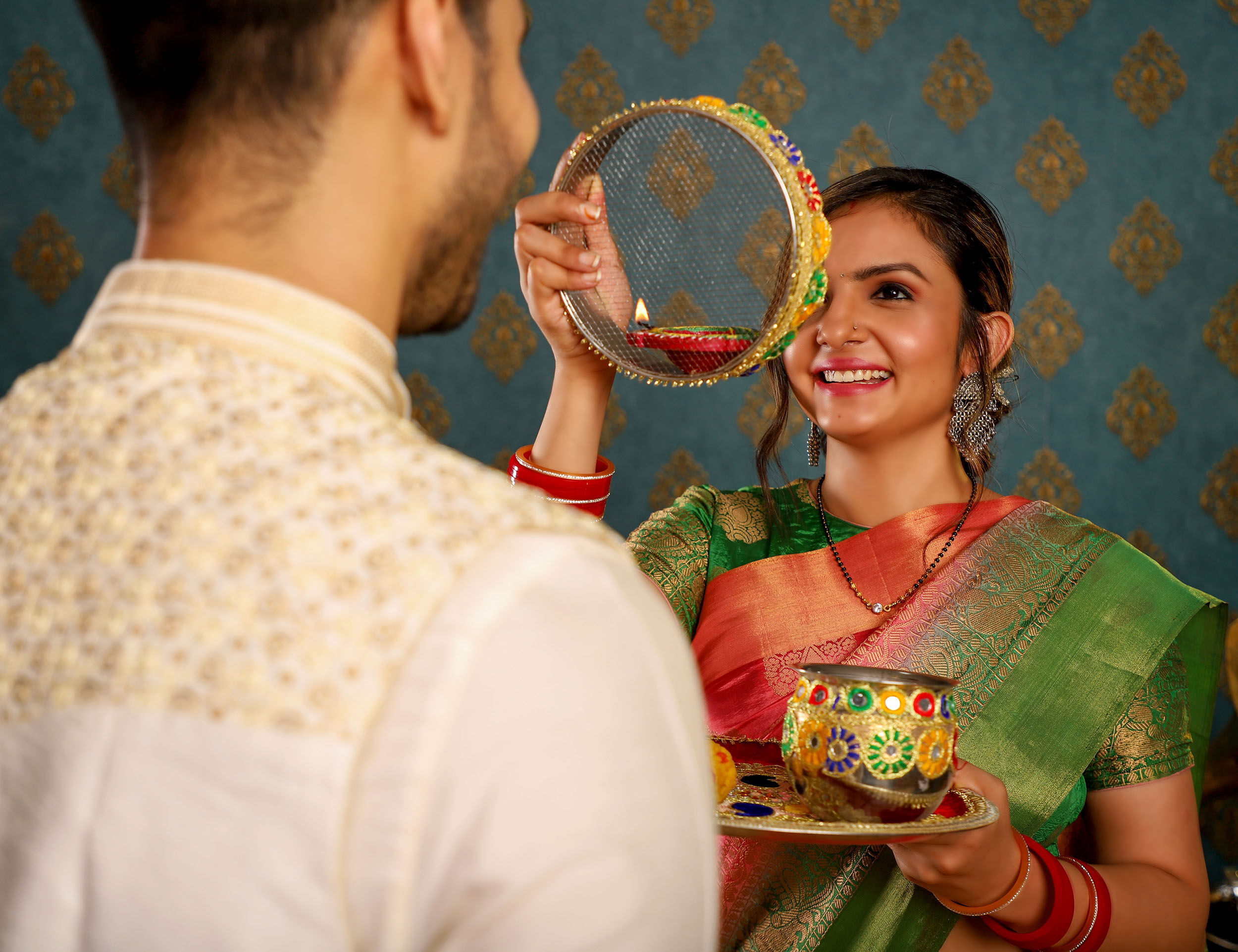 Woman performing Karwa Chauth ritual with decorated sieve and thali while looking at her husband